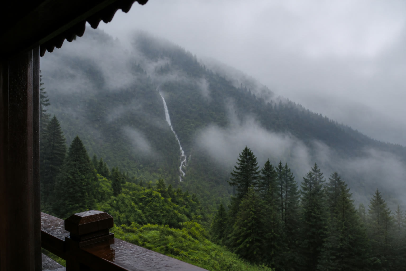 A waterfall and pine-covered Himalayan valley seen through a rain-darkened timber window